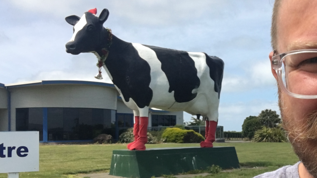 A selfie photo of a bearded man with glasses in front of a giant cow statue which has a Santa hat perched on its head and Christmas boots on its hooves. 