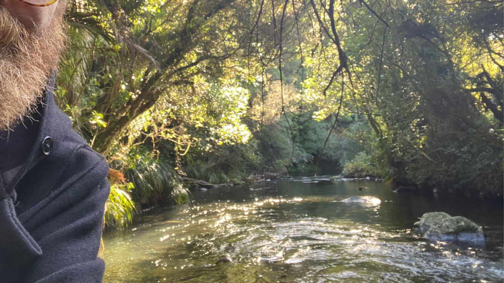 The side of a face of a bearded man in front of a river which is surrounded by dense native New Zealand bush which is beautifully lit.