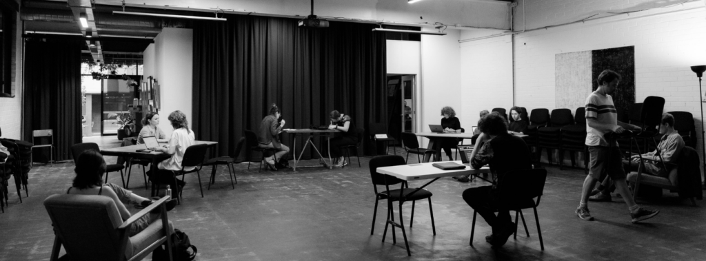 A black and white photo of writers sitting in the performance space of Tempo Rubato, they are writing.