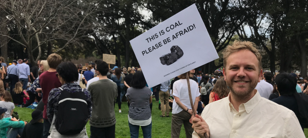 An image of an almost clean shaven JJW holding a sign which reads "This is coal. Please be afraid" with a picture of coal on it. He is at a packed Climate Strike protest.