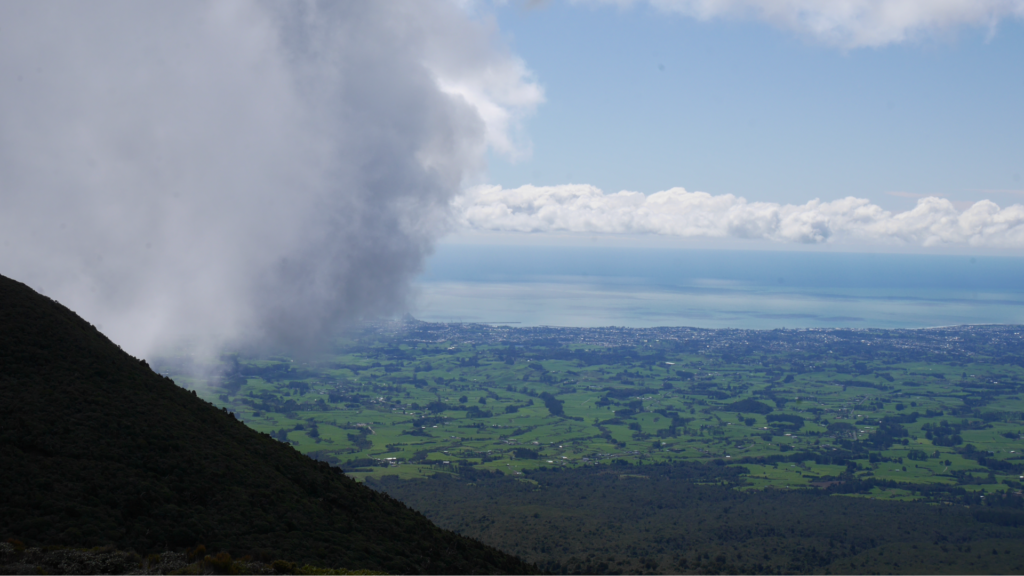 A fluffy cloud partially obscures a photo of New Plymouth taken from about 650m elevation up Mt Taranaki. You can see sprawling grassland from the edge of the National Park to the sea.