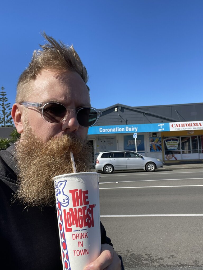 A weird looking blonde man with a large bead awkwardly slurps from a milkshake in a "THE LONGEST DRINK IN TOWN" cup.