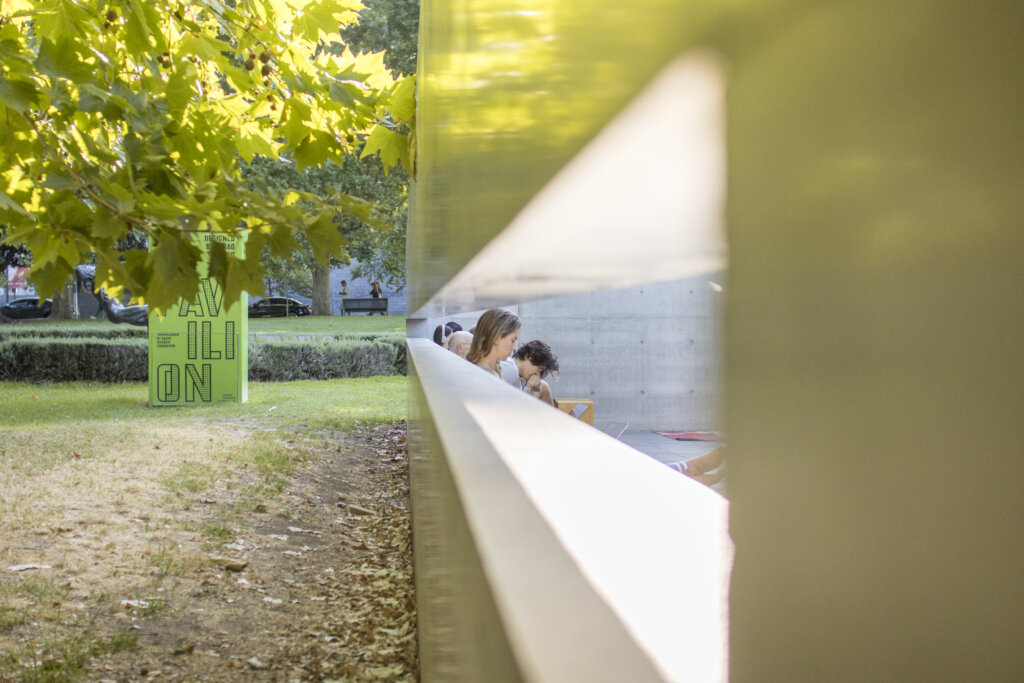 A smooth concrete wall reflects the warm summer evening light bouncing off a plane tree. A large green pillar with the MPavilion Logo on it. Writers writing visible through a gap in the concrete.